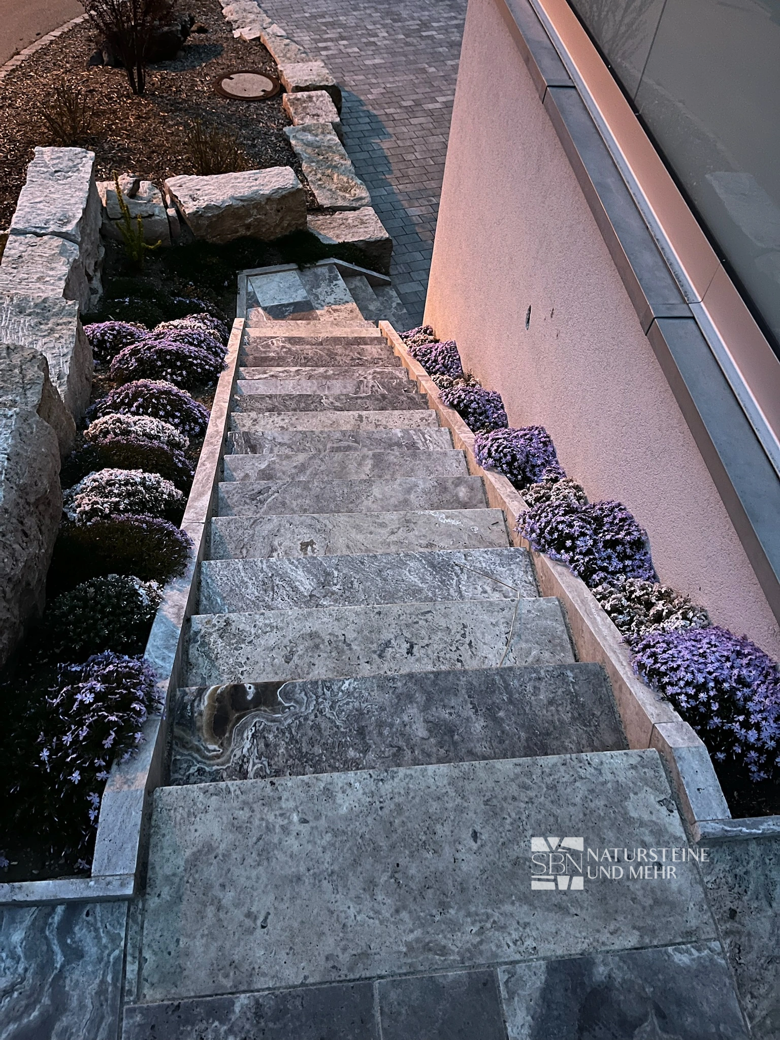 Schmale graubeige Natursteintreppe neben einer Hauswand mit violett blühenden Polsterpflanzen am Rand.