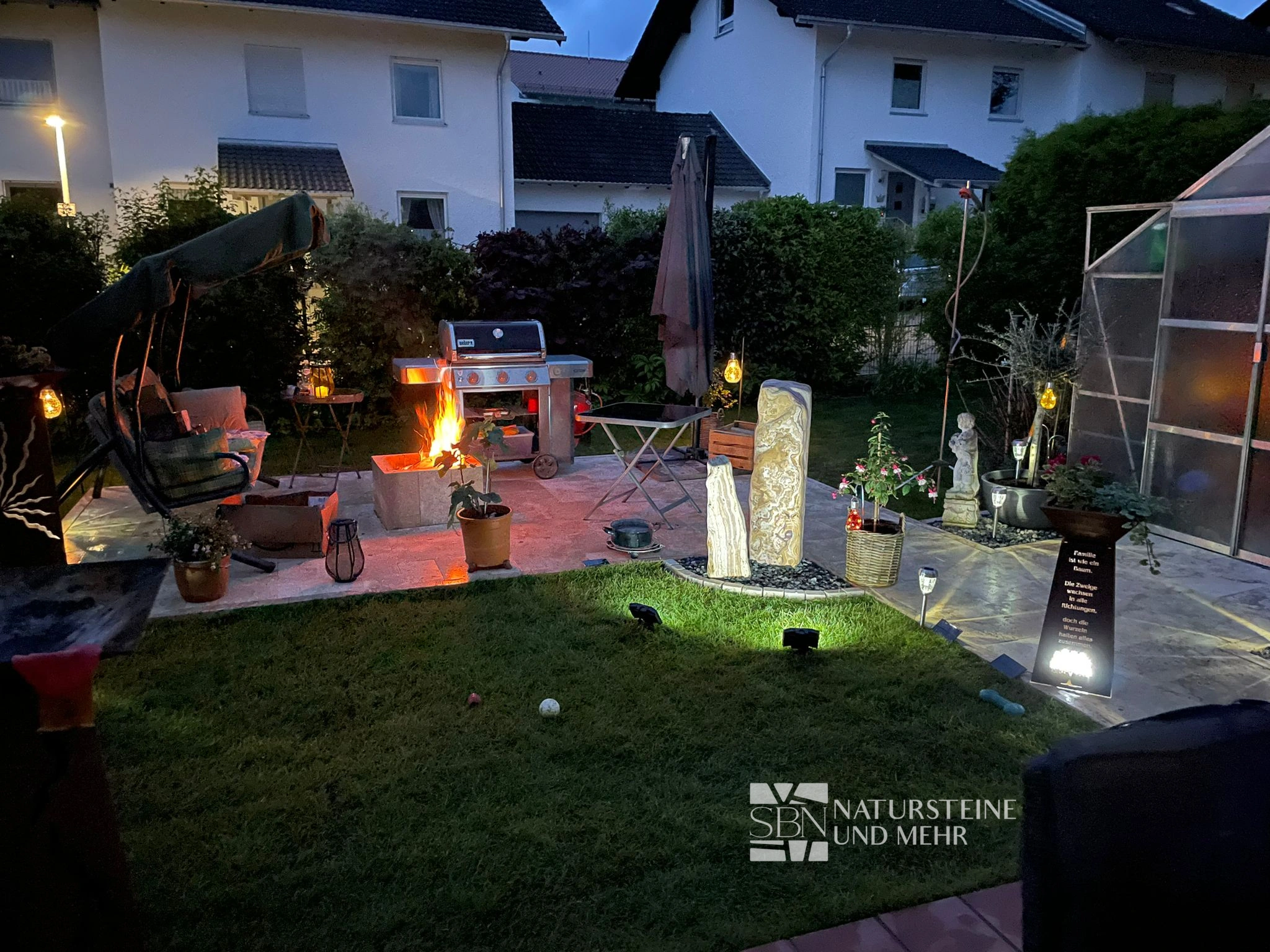 Travertinterrasse am Abend mit Feuerstelle, Sitzplatz, beleuchteten Gartendetails und eingefasster Rasenfläche.